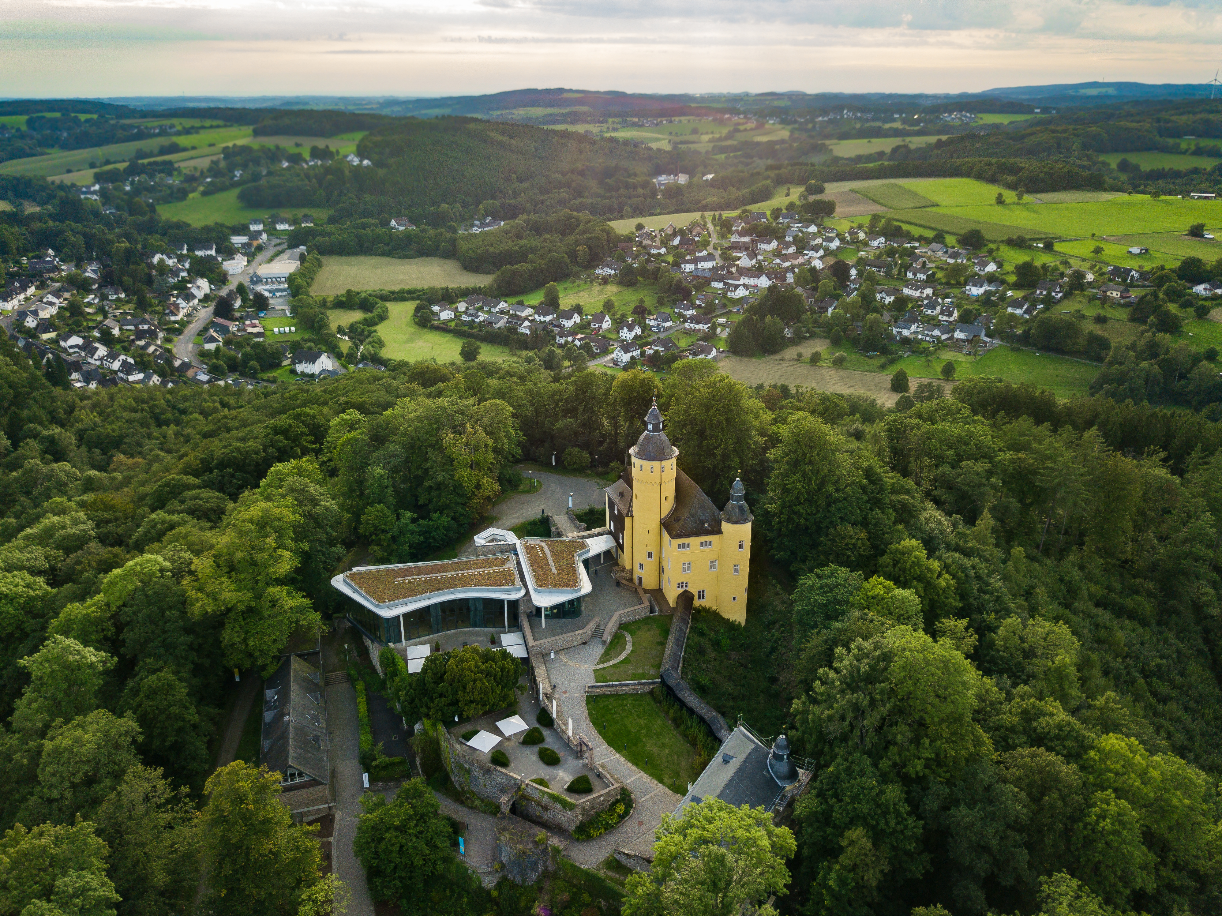 Luftaufnahme einer gelben Burg mit Türmen, umgeben von Bäumen. Im Vordergrund moderne Gebäude mit begrünten Dächern. Im Hintergrund eine ländliche Landschaft mit Feldern und Häusern.
