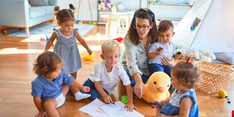 Beautiful teacher and group of toddlers sitting on the floor drawing using paper and pencil around lots of toys at kindergarten