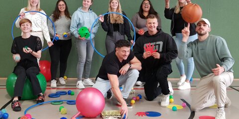 Eine Gruppe junger Menschen in einer Turnhalle posiert fröhlich mit bunten Sport- und Spielgeräten wie Reifen, Bällen und Würfeln auf dem Boden.