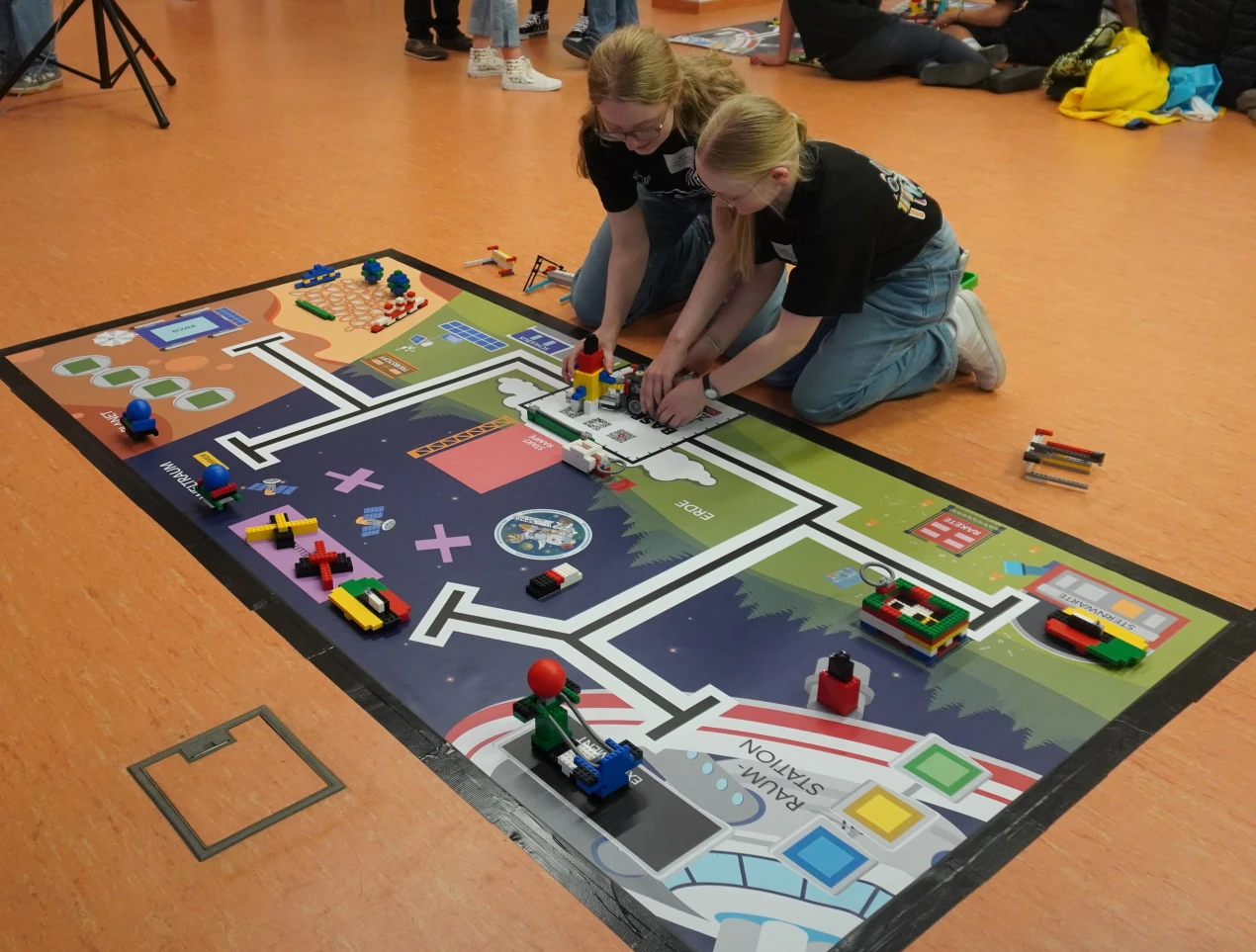 Zwei Personen arbeiten auf einem Boden an einem LEGO-Modell auf einer bedruckten Matte mit verschiedenen Bereichen und bunten LEGO-Gebilden. Weitere Menschen sind im Hintergrund sichtbar.