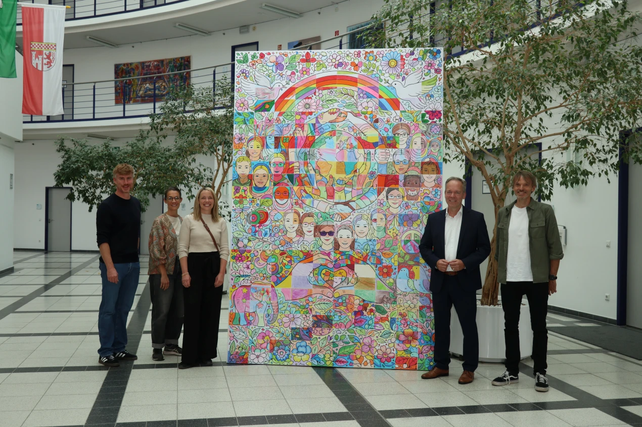 Fünf Personen stehen in einer modernen Halle vor einem großen, bunten Kunstwerk mit Motiven wie Regenbögen, Blumen, Tauben und Gesichtern. Im Hintergrund sind Pflanzen, Flaggen und Türen zu sehen.