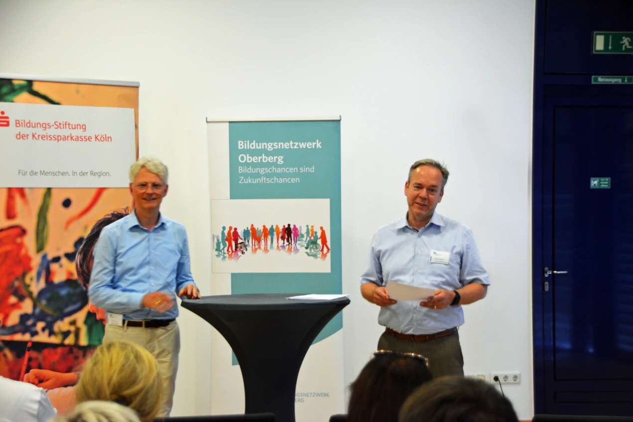 Zwei Männer stehen an einem Stehtisch vor Bannern von Bildungsnetzwerk Oberberg und Bildungs-Stiftung der Kreissparkasse Köln. Im Vordergrund sitzen Personen.