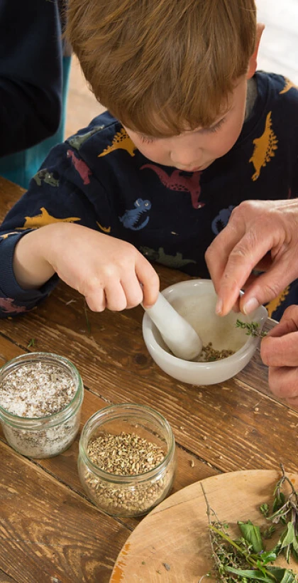 Zwei Kinder und eine erwachsene Person bereiten frische Kräuter an einem Holztisch vor. Ein Kind nutzt einen Mörser, während das andere Kräuter hackt.