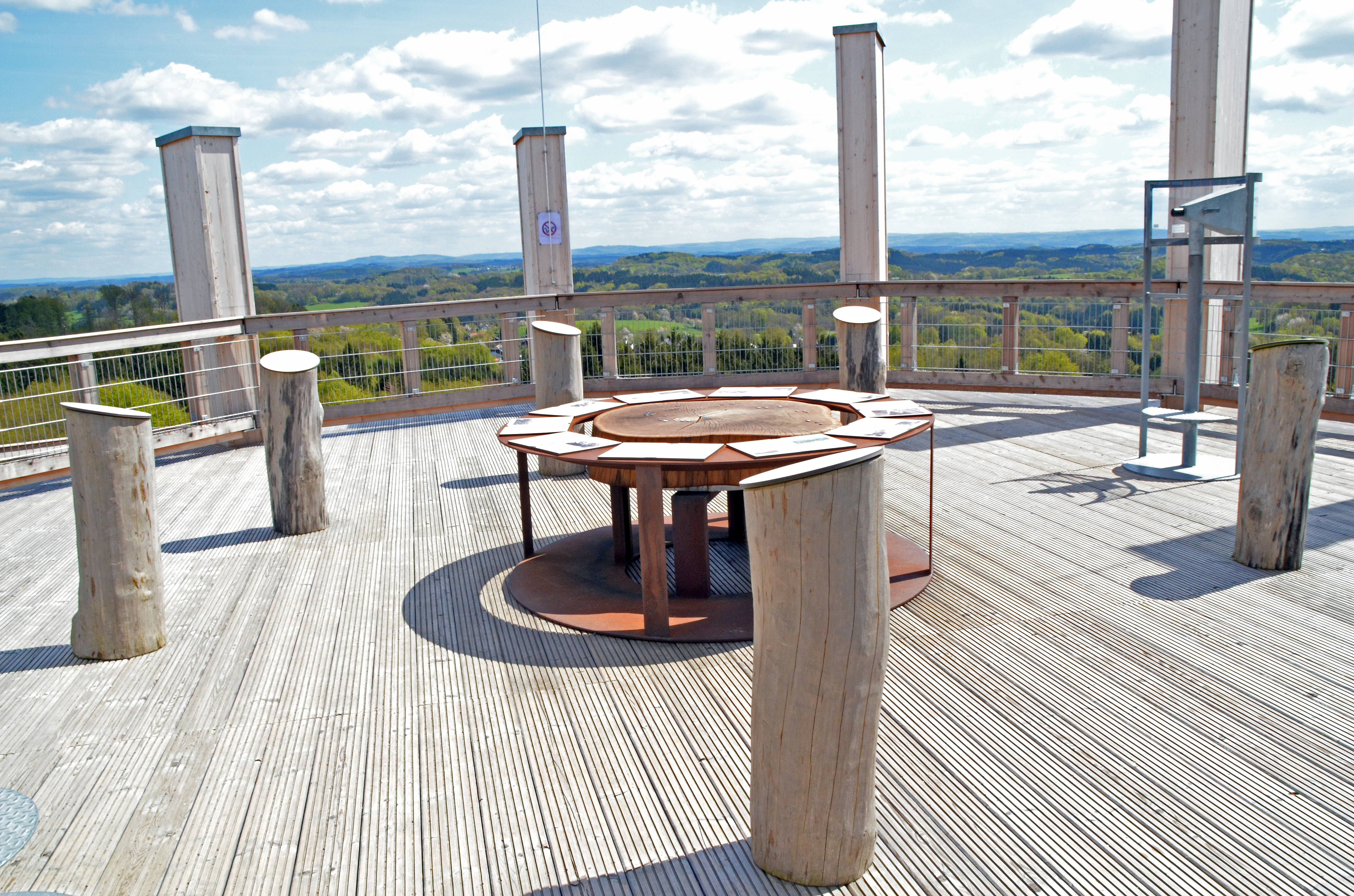 Aussichtsplattform mit Holzsäulen, einem runden Metalltisch in der Mitte und weitläufigem Blick auf eine grüne, hügelige Landschaft unter blauem Himmel.