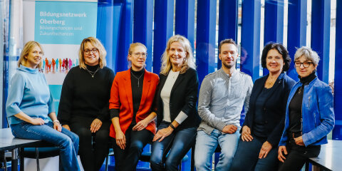 Eine Gruppe von sieben Personen sitzt nebeneinander vor einem Hintergrund mit blauen vertikalen Streifen. Links steht ein Banner mit der Aufschrift 'Bildungsnetzwerk Oberberg - Bildungschancen sind Zukunftschancen'.
