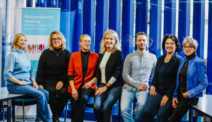 Eine Gruppe von sieben Personen sitzt nebeneinander vor einem Hintergrund mit blauen vertikalen Streifen. Links steht ein Banner mit der Aufschrift 'Bildungsnetzwerk Oberberg - Bildungschancen sind Zukunftschancen'.