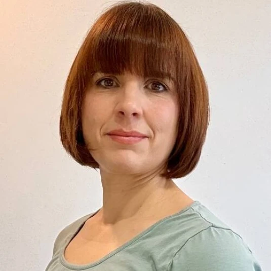 Eine Frau mit kinnlangem braunem Haar und Pony trägt ein hellgrünes Oberteil. Sie schaut in die Kamera vor einem neutralen, hellen Hintergrund.