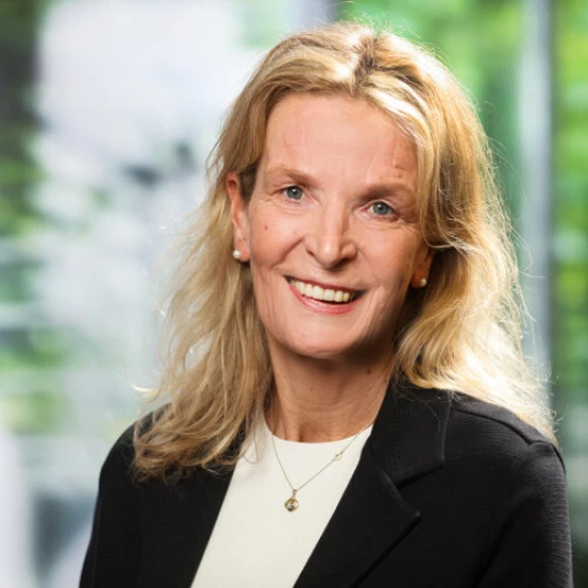 Porträt einer lächelnden Frau mit blonden Haaren, schwarzem Blazer und hellem Oberteil. Unscharfer Hintergrund mit grünen und weißen Farben.