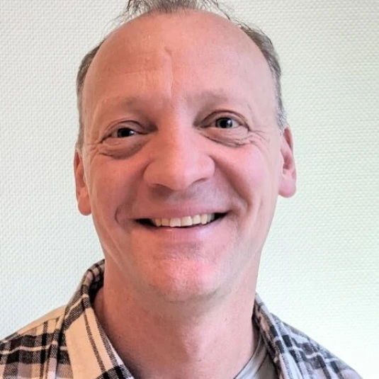 Porträt einer lächelnden Person mit langen blonden Haaren, die wellig herabfallen. Die Person trägt ein dunkles Oberteil vor einem unscharfen Hintergrund.