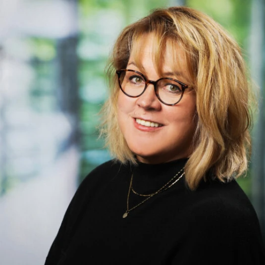 Porträt einer lächelnden Person mit blonden schulterlangen Haaren und Brille. Die Person trägt einen schwarzen Pullover und eine goldene Halskette. Der Hintergrund ist unscharf und zeigt grüne und weiße Farbtöne.